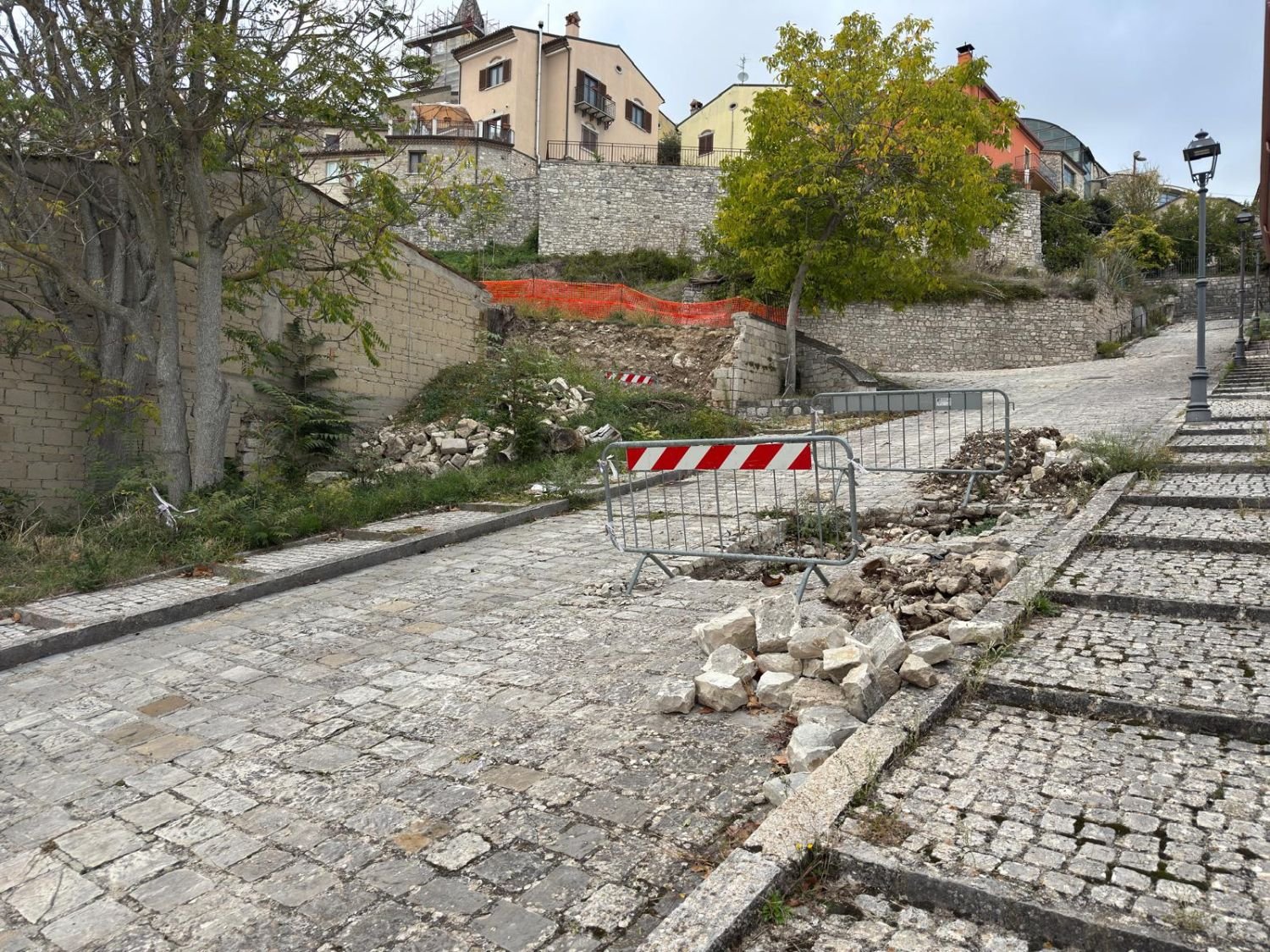 Tratto di muro crollato con rete arancione, pietre accatastate e strada transennata